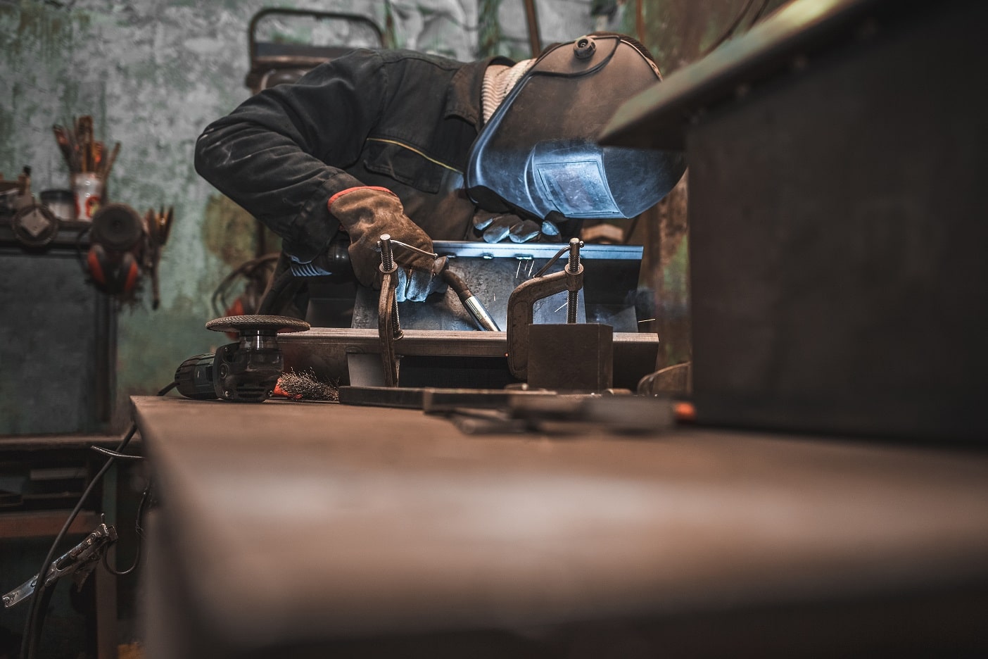 A worker in welder mask does MIG welding in a workshop.
