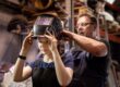 A welding mentor helps his apprentice to put on her welding mask.