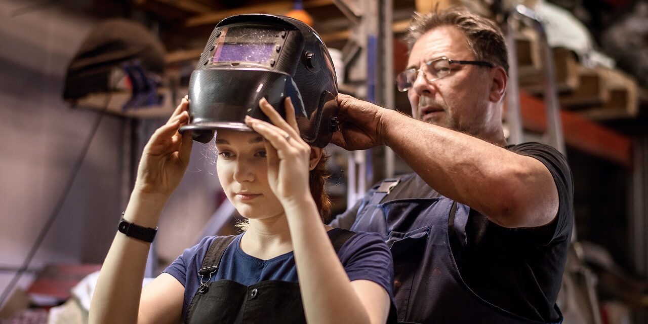 welder-mentor-helps-apprentice-put-on-welding-mask A welding mentor helps his apprentice to put on her welding mask.