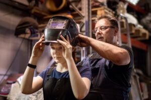A welding mentor helps his apprentice to put on her welding mask.
