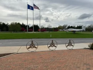 A custom welded metal installation of paver guards with an interior propeller design sits in front of a sidewalk, road, and grass where a fighter jet sits.