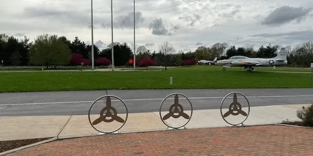 Air Zoo Custom Welded Metal Installations-min A custom welded metal installation of paver guards with an interior propeller design sits in front of a sidewalk, road, and grass where a fighter jet sits.