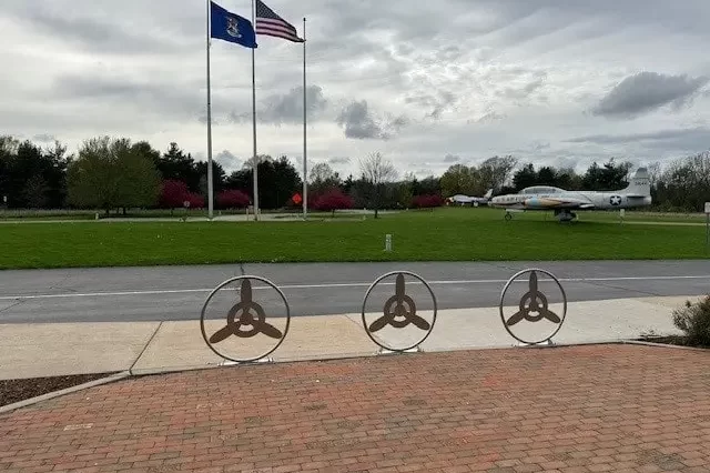 Air Zoo Custom Welded Metal Installations-min A custom welded metal installation of paver guards with an interior propeller design sits in front of a sidewalk, road, and grass where a fighter jet sits.