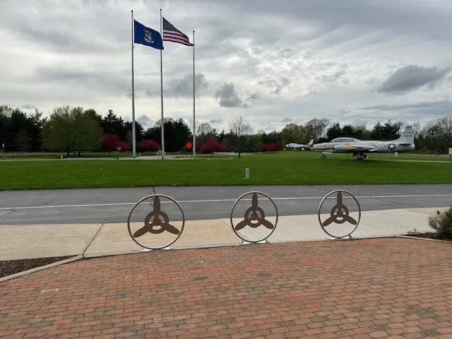 Air Zoo Custom Welded Metal Installations-min A custom welded metal installation of paver guards with an interior propeller design sits in front of a sidewalk, road, and grass where a fighter jet sits.
