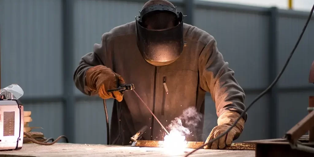 the-man-works-with-a-welding-machine-2023-11-27-05-24-32-utc-min A man wearing a jumpsuit, welding mask, and gloves, works with a welding machine outdoors.