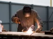 A man wearing a jumpsuit, welding mask, and gloves, works with a welding machine outdoors.