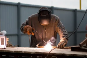 A man wearing a jumpsuit, welding mask, and gloves, works with a welding machine outdoors.