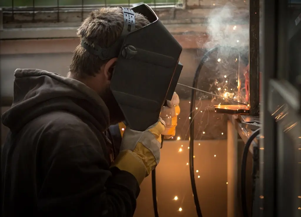 A young man with a welding mask and tool performs welding repairs on a metal structure.