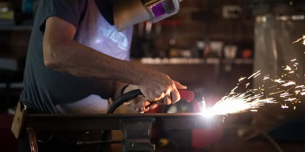 welding-2023-11-27-04-58-45-utc-min A man in a tee shirt and welding mask performs a weld on a piece of metal.