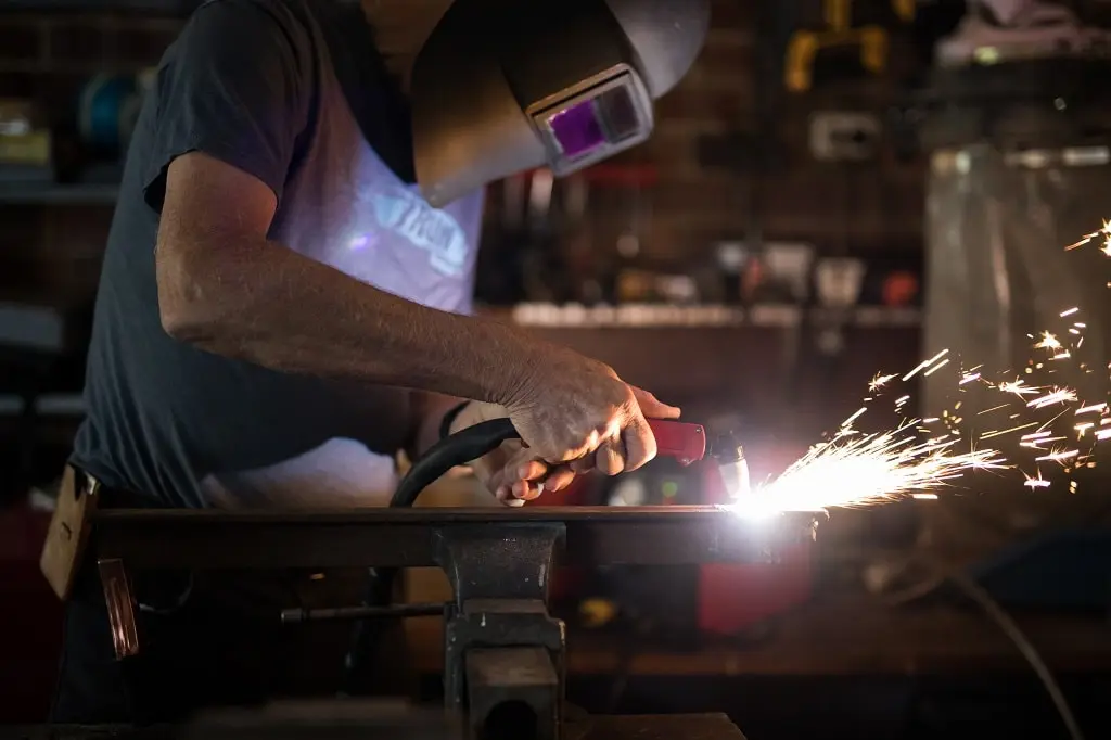 A man in a tee shirt and welding mask performs a weld on a piece of metal.
