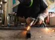 A welder in a protective helmet welds together two metal bars.