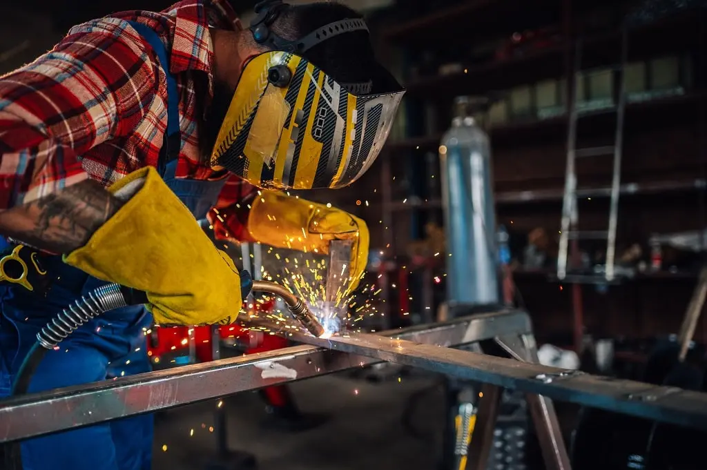 Skilled worker in protective gear performs metal fabrication by welding two metal rods together.