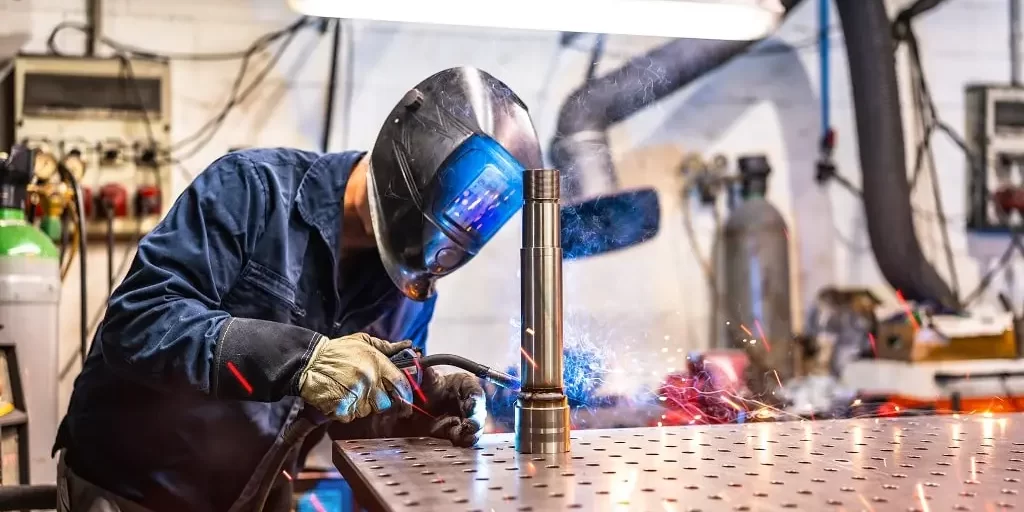welder-with-safety-work-wear-working-in-factory-2024-12-10-03-23-59-utc-min A welder with a safety mask on works in a factory, welding a metal tube to another component part.