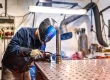 A welder with a safety mask on works in a factory, welding a metal tube to another component part.