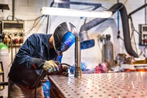 A welder with a safety mask on works in a factory, welding a metal tube to another component part.